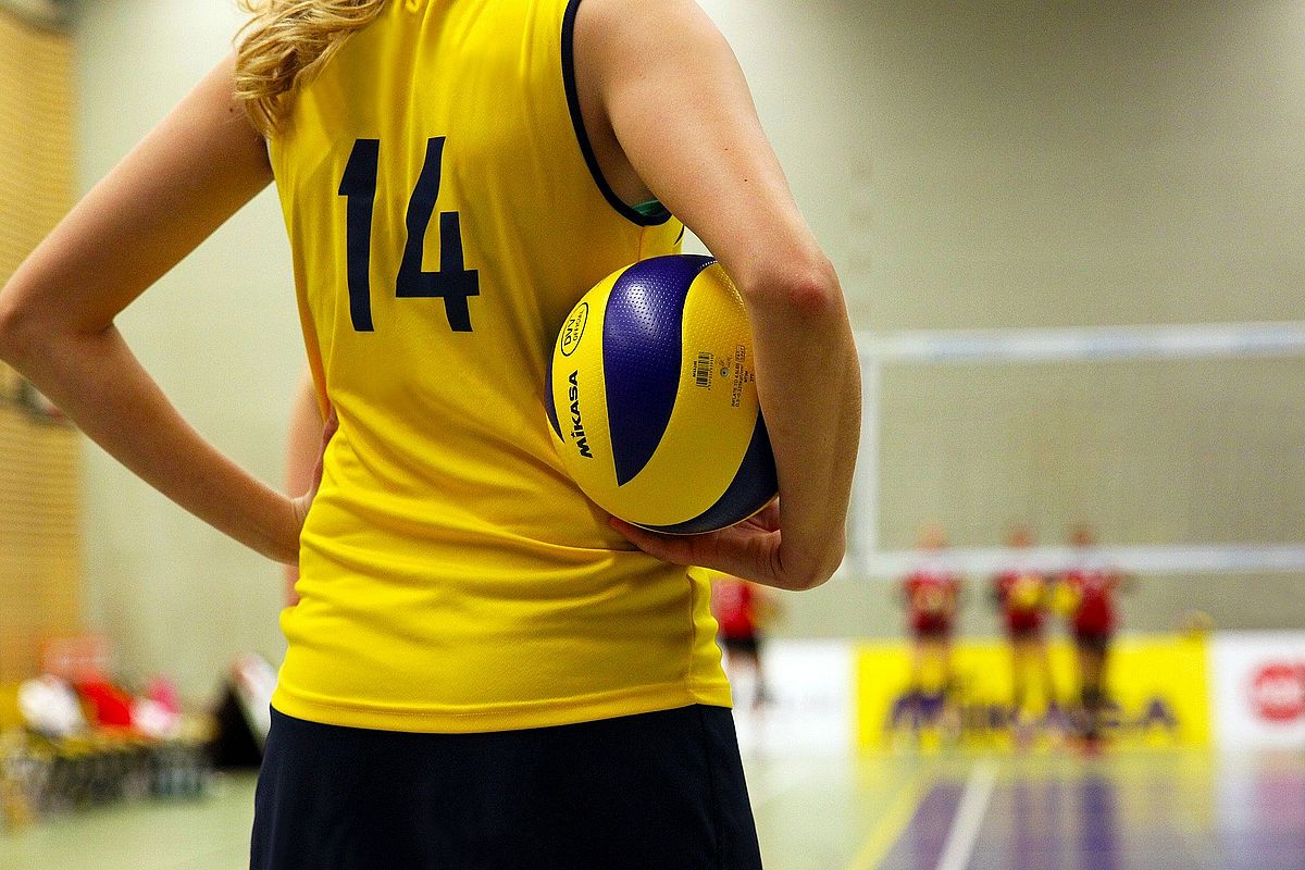 eine Frau steht in einer Sporthalle in einem gelben ärmellosen Dress mit einem Volleyball in der Hand. Man sieht nur ihren Rücken mit der Nummer 14